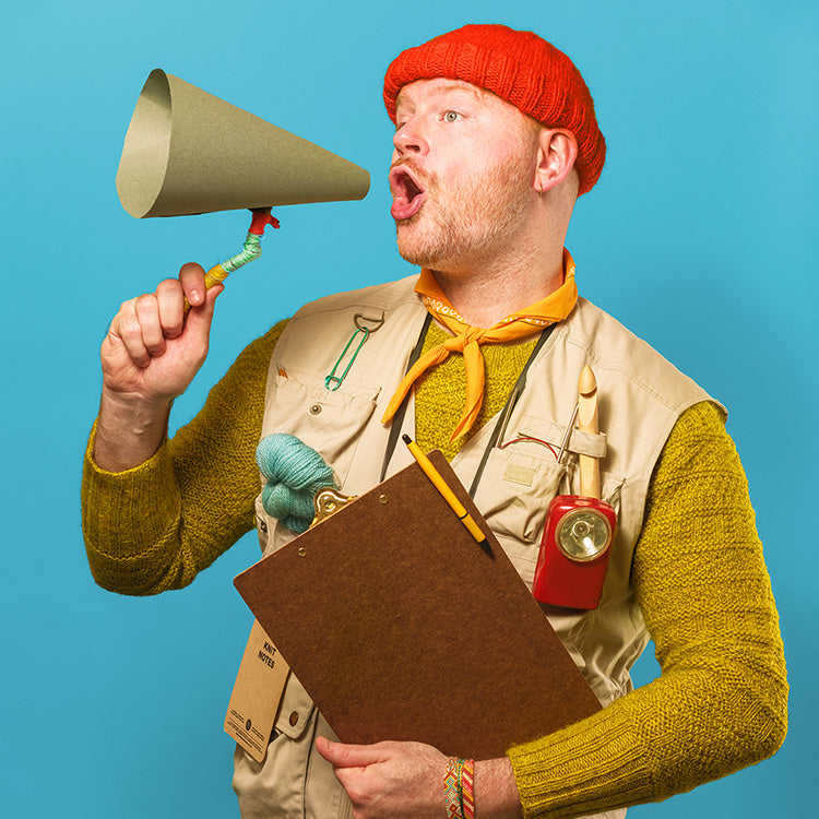 The image shows Stephen West dressed in camping core, using a handmade megaphone. The background is bright blue.  
