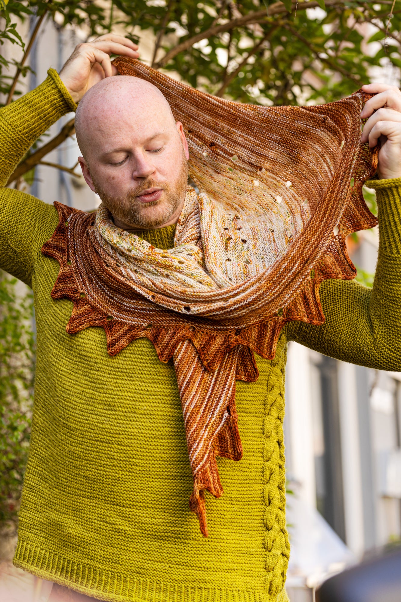 This image shows Stephen West wearing a beautifully knitted sample of the Camp Craft Shawl by Westknits, part of the 2025 YAL. The sample is in warm, brown tones. 