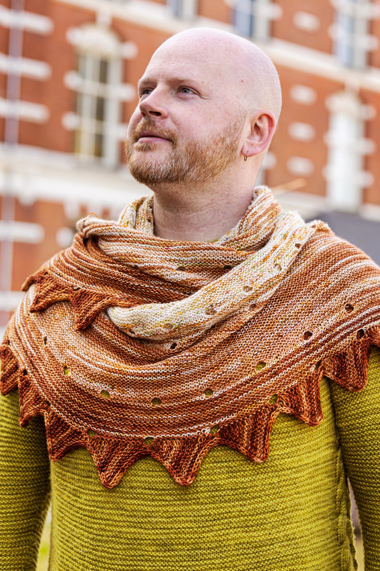 This image shows Stephen West wearing a beautifully knitted sample of the Camp Craft Shawl by Westknits, part of the 2025 YAL. The sample is in warm, brown tones. 