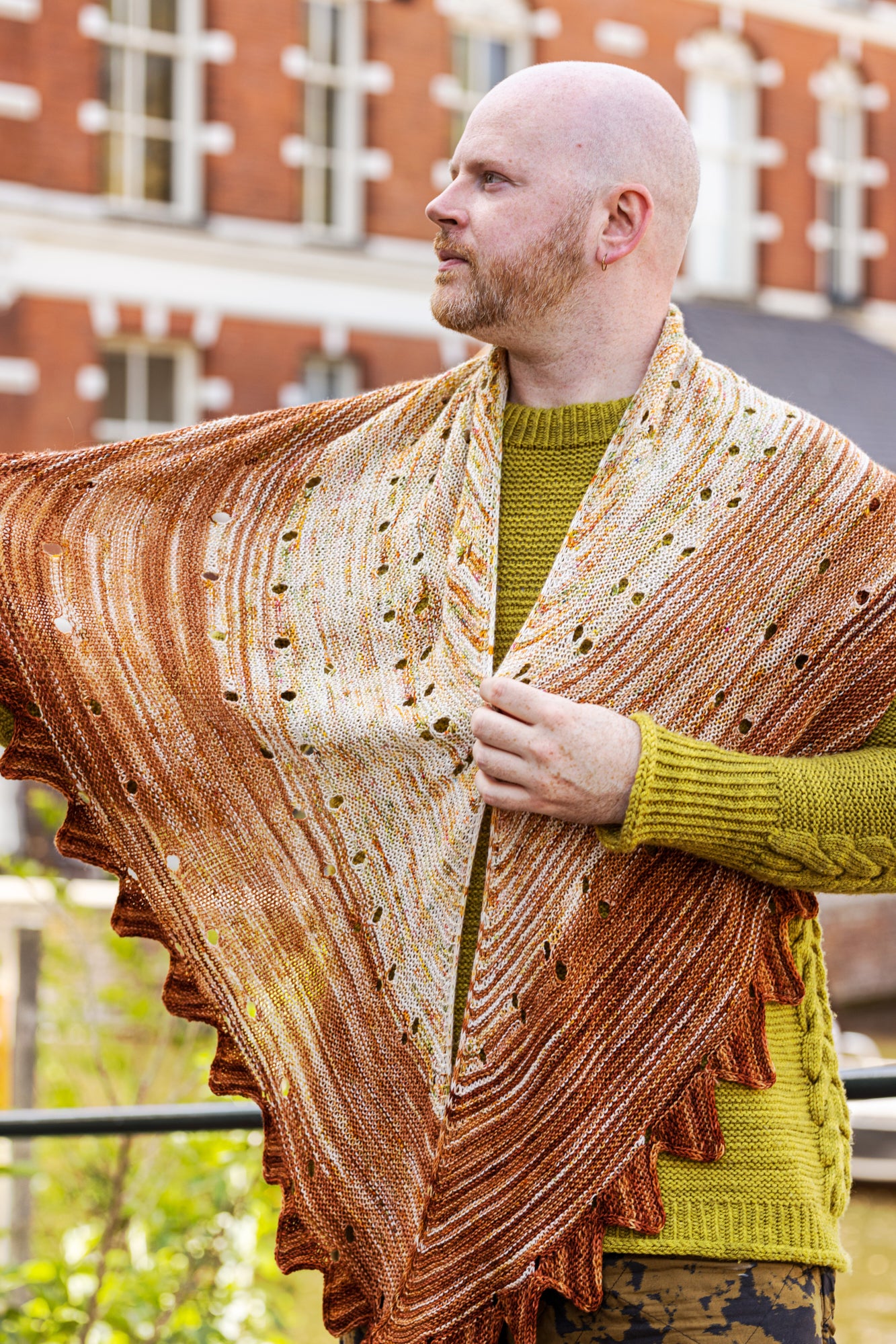 This image shows Stephen West wearing a beautifully knitted sample of the Camp Craft Shawl by Westknits, part of the 2025 YAL. The sample is in warm, brown tones. 