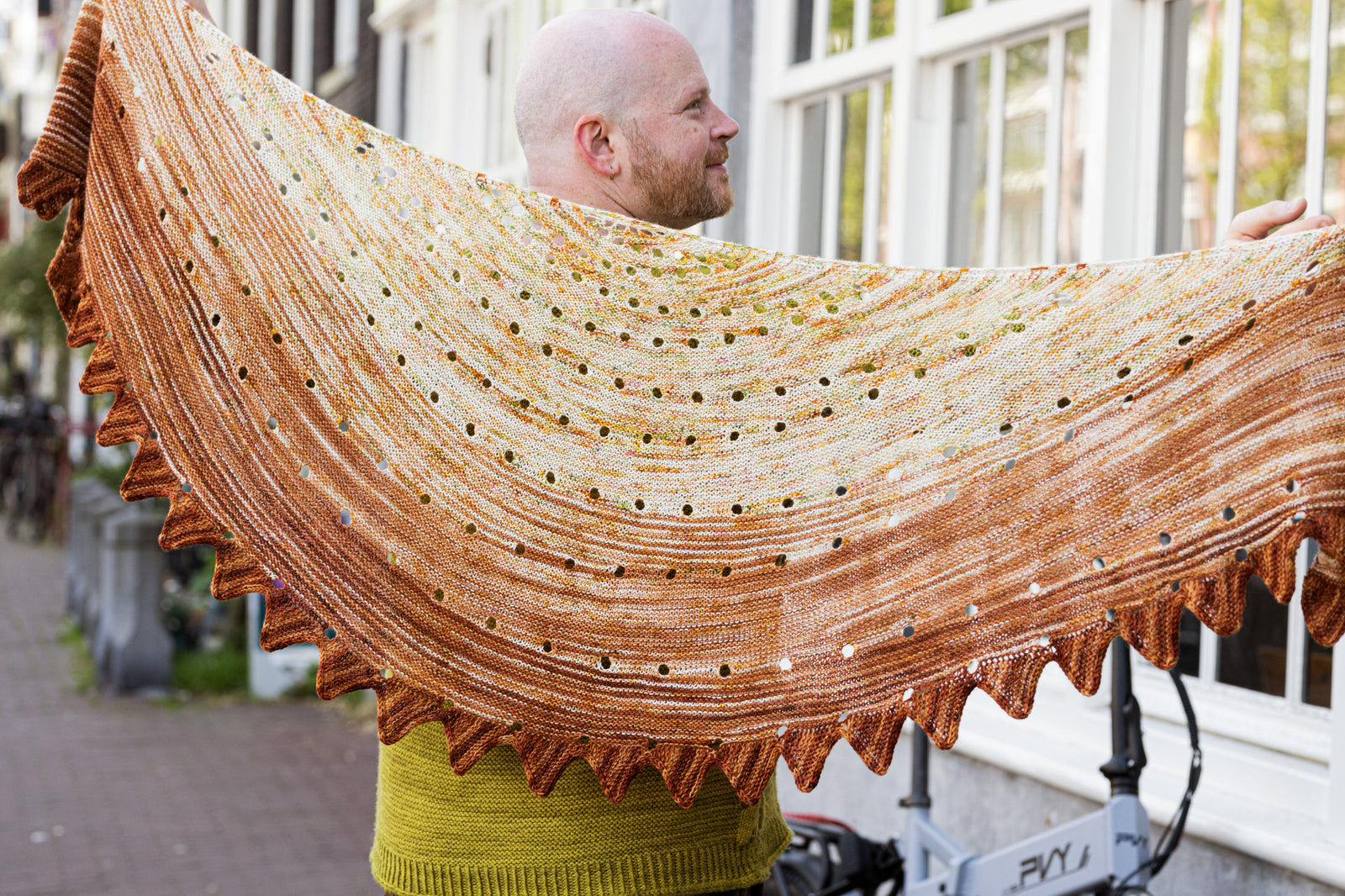 This image shows Stephen West holding a beautifully knitted sample of the Camp Craft Shawl by Westknits, part of the 2025 YAL. The sample is in warm, brown tones. 