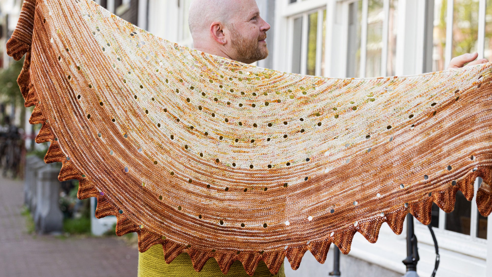 This image shows Stephen West holding a beautifully knitted sample of the Camp Craft Shawl by Westknits, part of the 2025 YAL. The sample is in warm, brown tones. 