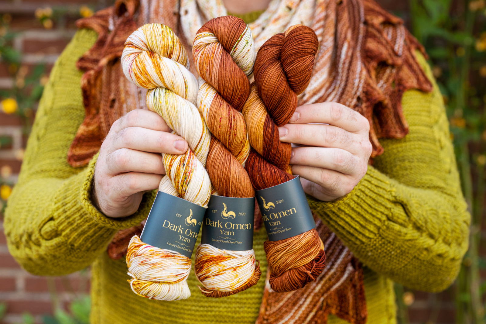 This image shows Stephen West wearing a beautifully knitted sample of the Camp Craft Shawl by Westknits, part of the 2025 YAL. The sample is in warm, brown tones. He holds three skeins of Dark Omen Yarn in brown tones. 