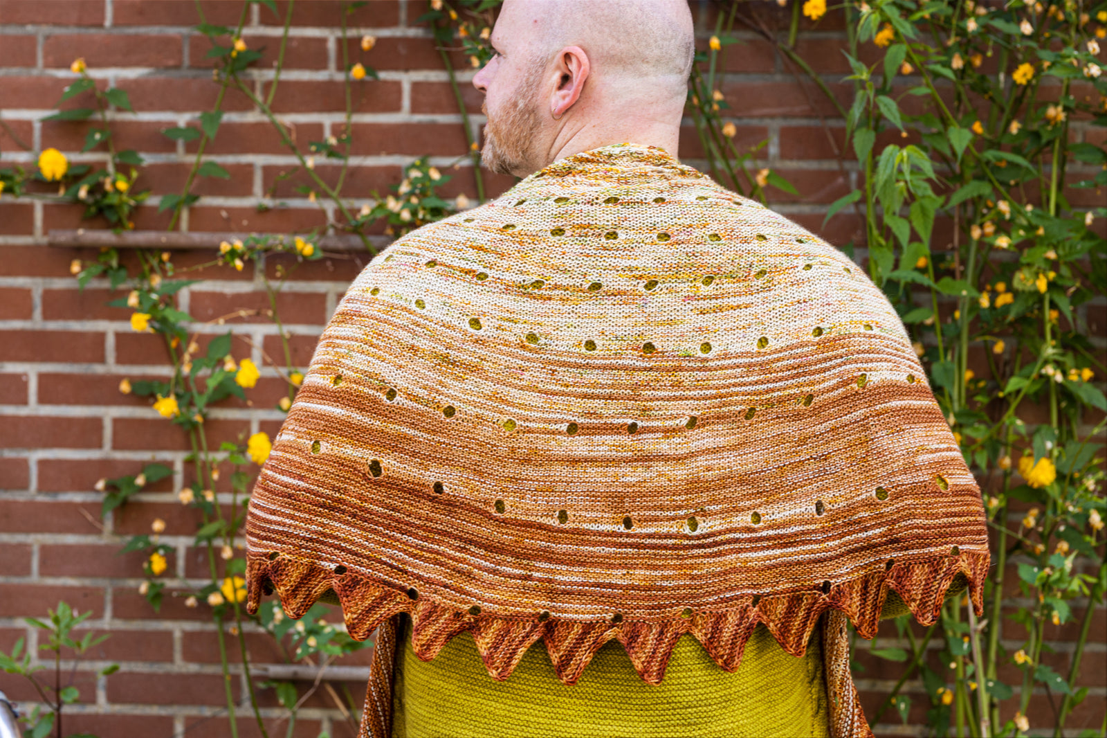 This image shows Stephen West wearing a beautifully knitted sample of the Camp Craft Shawl by Westknits, part of the 2025 YAL. The sample is in warm, brown tones. 