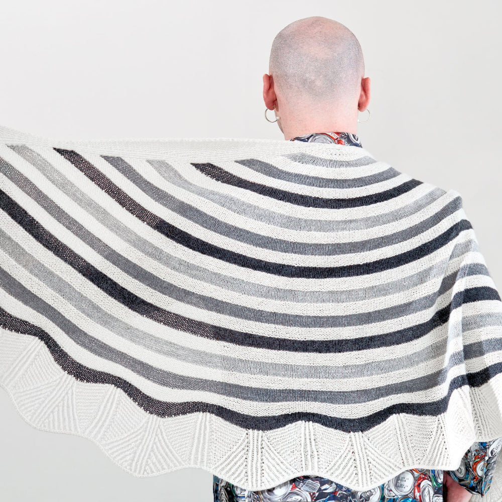 This image shows Stephen West wearing a beautifully knitted sample of the Canal Ring Shawl from Westknits in contrasting grey and white tones. 