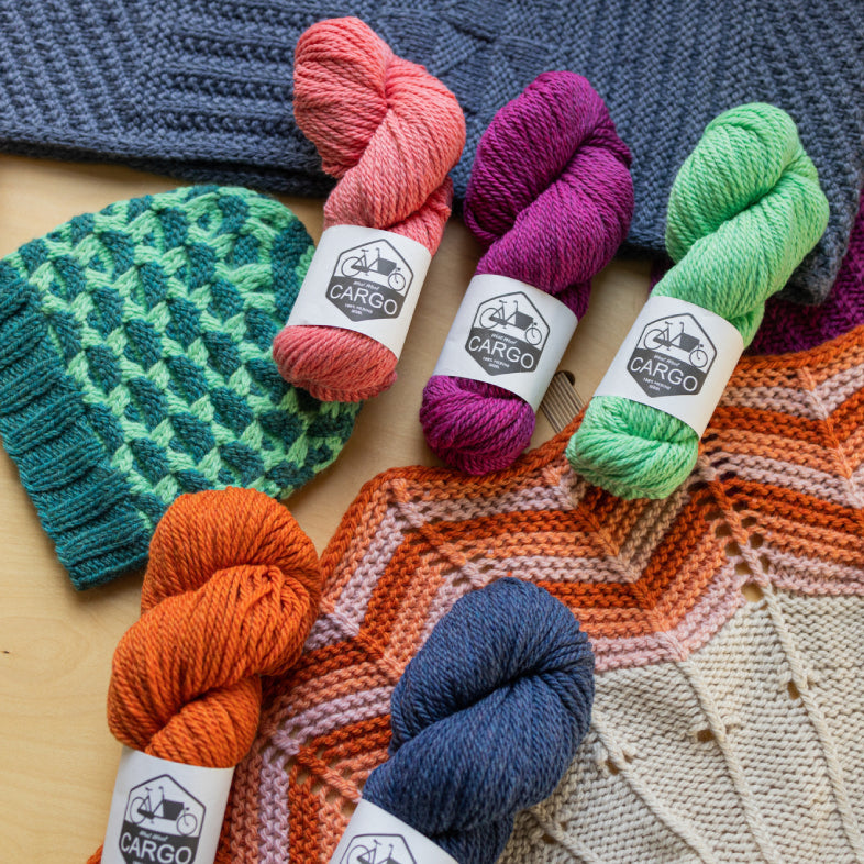 This image shows various skeins of chunky weight Cargo  from West Wool  in various colours, such as coral, green, blue, and purple. They are laid on various knitted samples. 