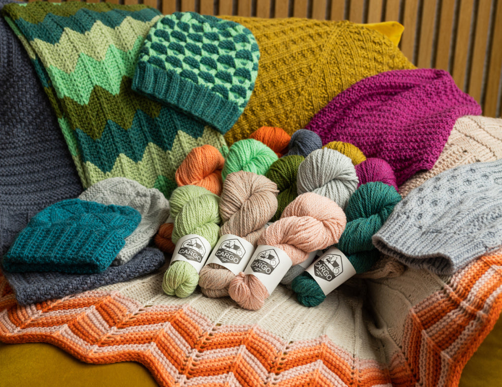 This image shows various skeins of chunky weight Cargo from West Wool in various colours, such as coral, green, blue, and purple. They are laid on various knitted samples. 