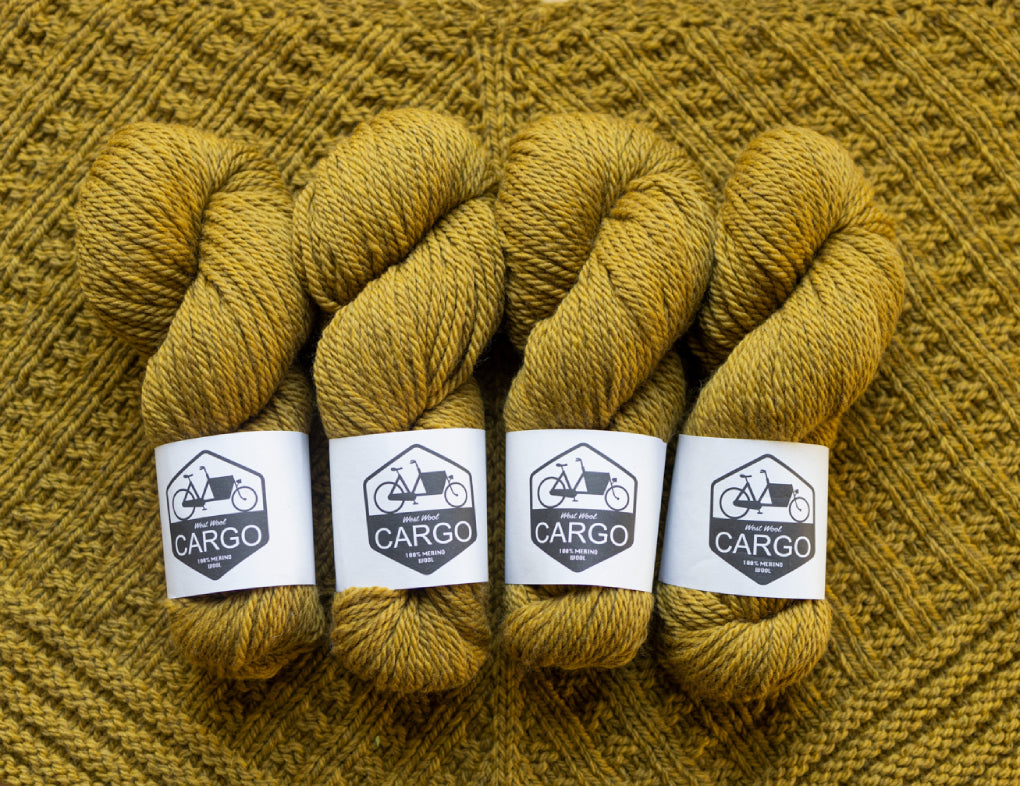 This image shows four skeins of West Wool Cargo in ochre, laid on a beautifully knitted shawl in the same colour. 