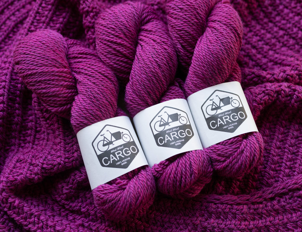 This image shows three skeins of West Wool Cargo in fuchsia, laid on a beautifully knitted shawl in the same colour. 