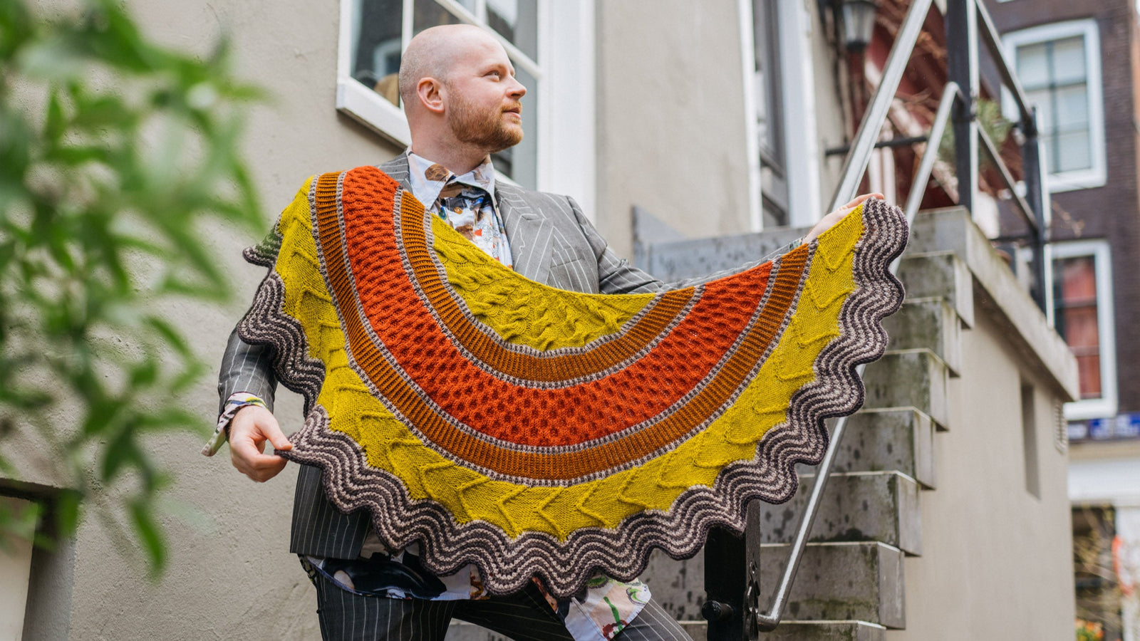 This image shows Stephen West wearing a beautifully knitted sample of the Corrie Shawl Sample by Westknits in orange, yellow and brown tones. 