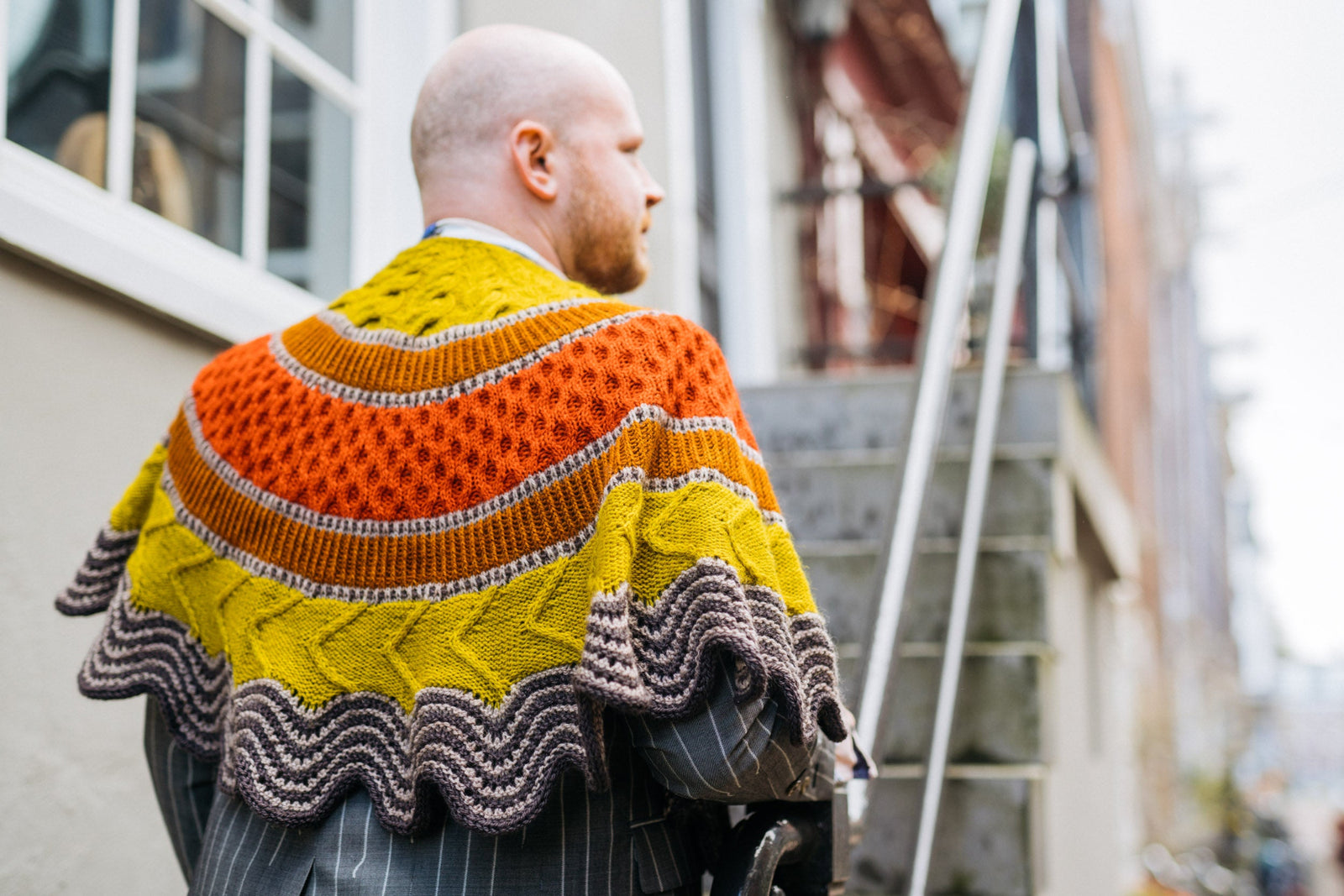 This image shows Stephen West wearing a beautifully knitted sample of the Corrie Shawl Sample by Westknits in orange, yellow and brown tones. 