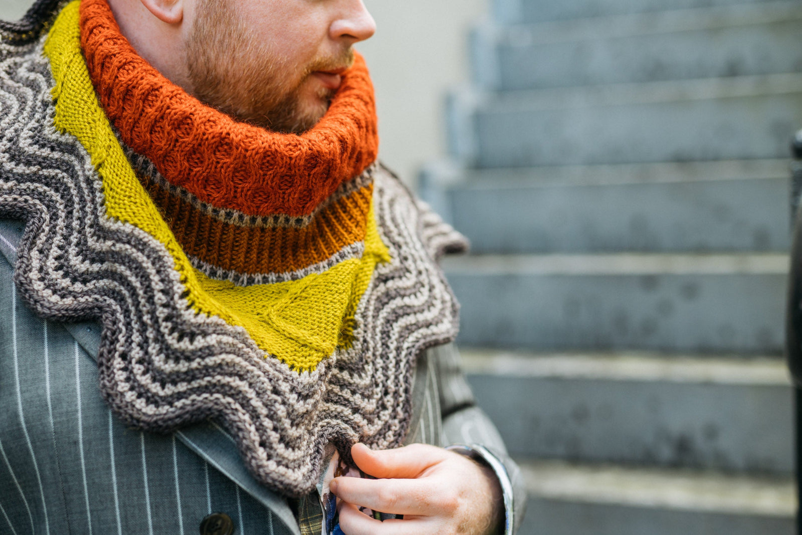 This image shows Stephen West wearing a beautifully knitted sample of the Corrie Shawl Sample by Westknits in orange, yellow and brown tones. 