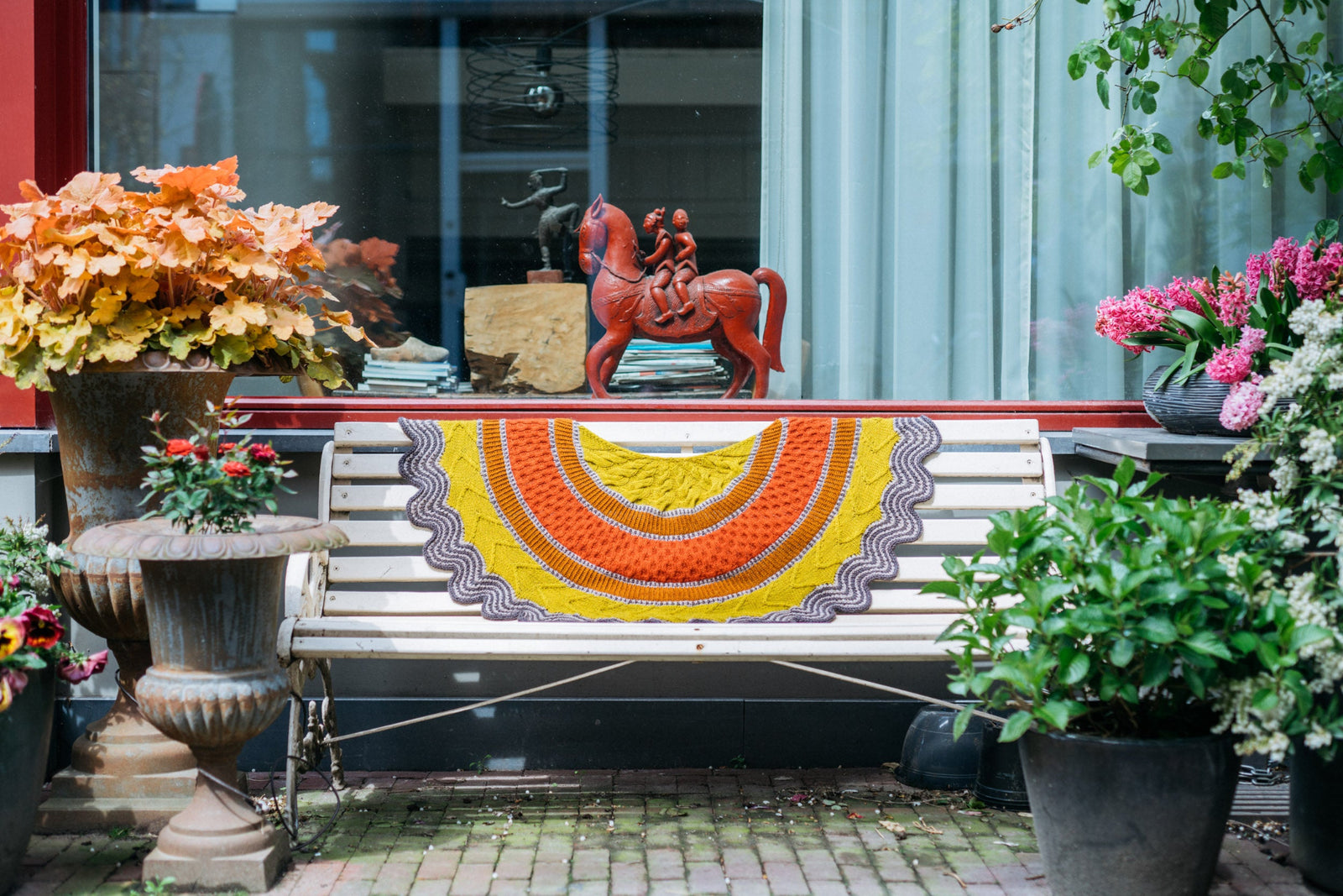 This image shows a beautifully knitted sample of the Corrie Shawl Sample by Westknits in orange, yellow and brown tones laid on a bench. 