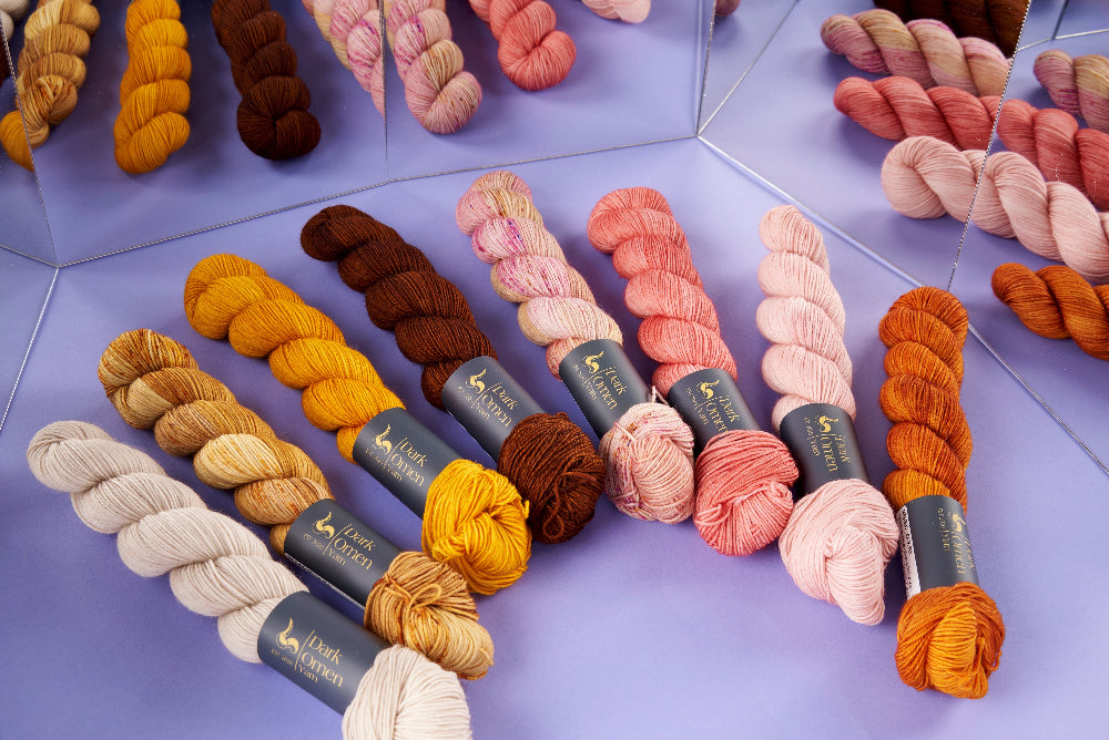 Multiple skeins of yarn in assorted colors arranged on a lavender surface with mirrored panels in the background. The yarns include cream, tan, mustard, brown, pink, peach, coral, and orange tones.