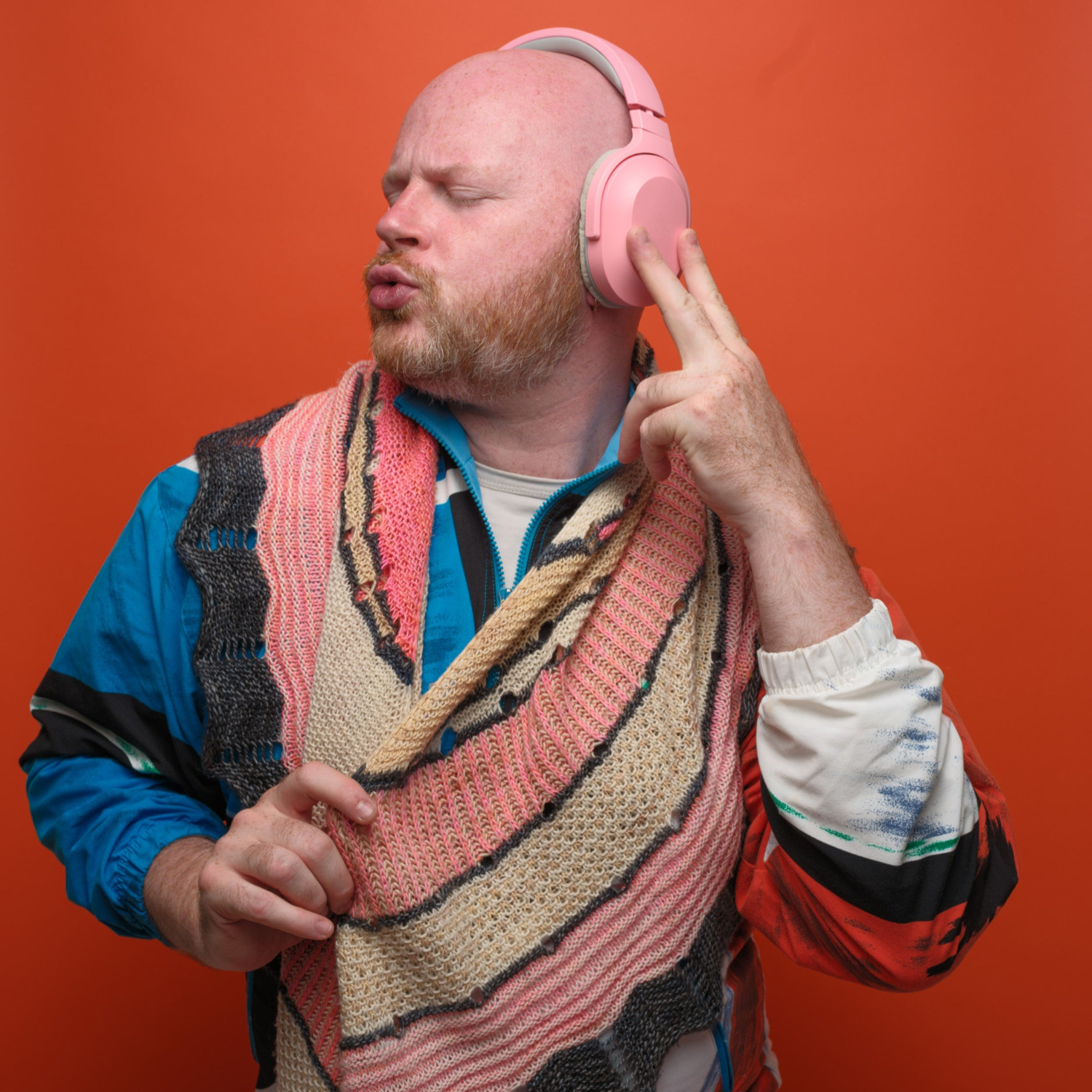 Stephen West wearing pink headphones and a colourful swawl against an orange background with a retro early 90s vibe