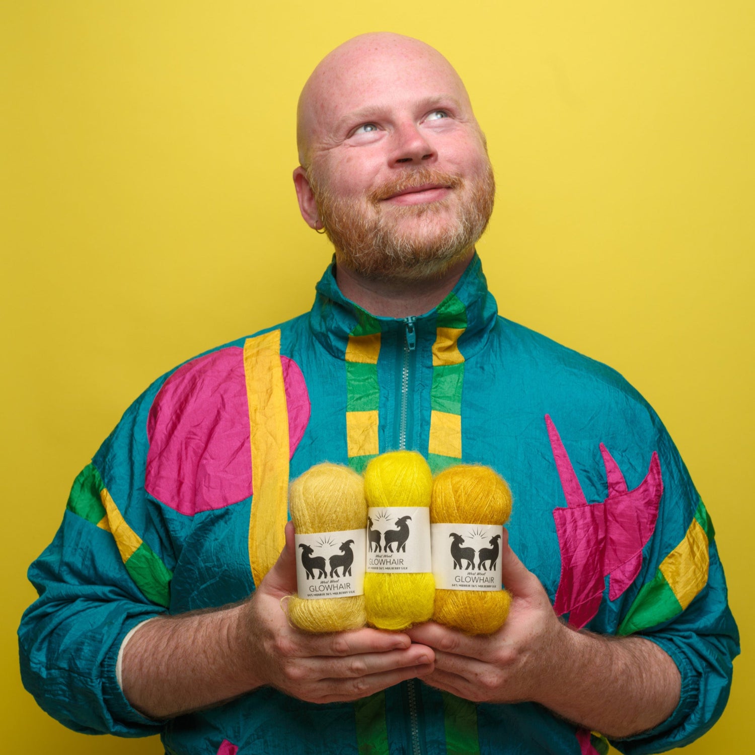 Stephen West wearing a colourful jacket holding three skeins of Glowhair lace weight yarn against a yellow background