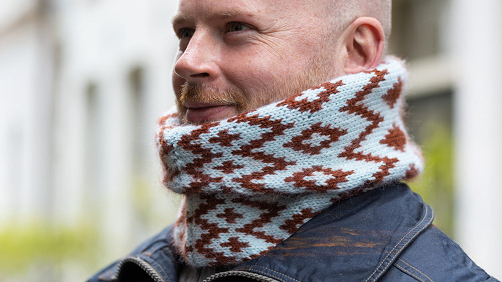 Stephen West wearing a knitted neck cowl in a diamond pattern in brown and light blue. Standing against a blurred background of buildings and greenery. 