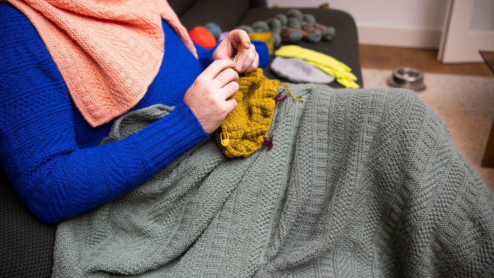 This image shows Stephen West knitting while wearing a knitted sample of the Dustland Blanket by Westknits in dusty green yarn. 