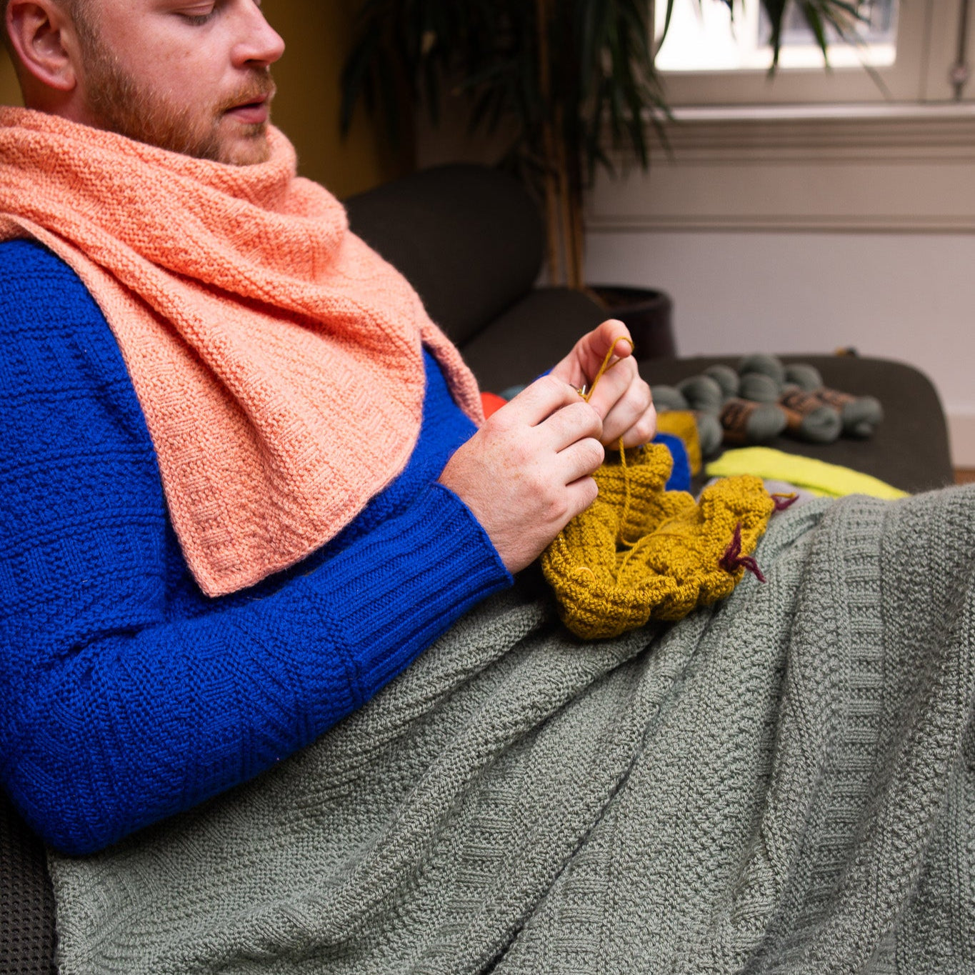 This image shows Stephen West knitting while wearing a knitted sample of the Dustland Blanket by Westknits in dusty green yarn. 
