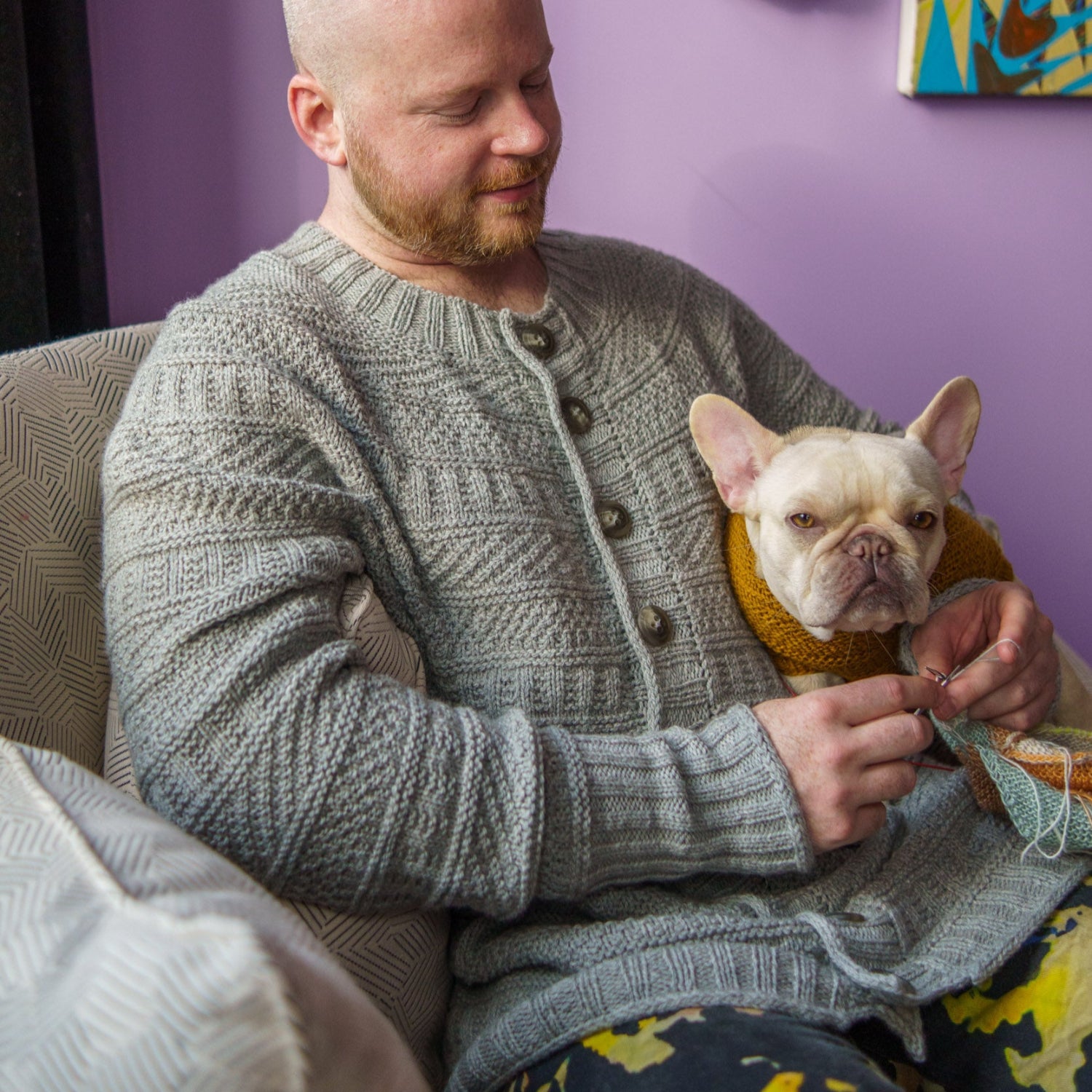 This image shows Stephen West wearing a beautifully knitted sample of the Dustland Cardigan in grey. Next to him, there is his dog. 