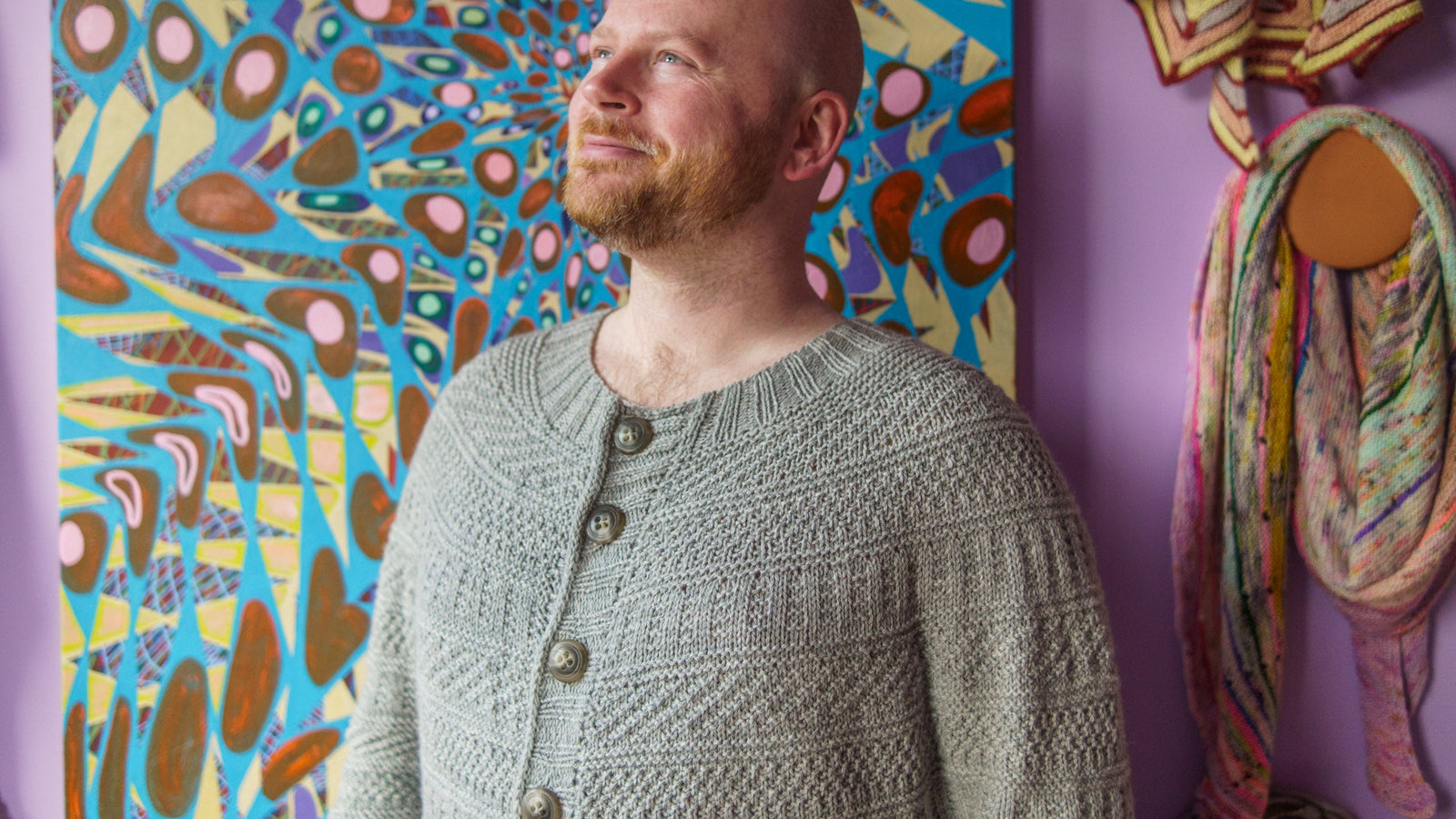 This image shows Stephen West wearing a beautifully knitted sample of the Dustland Cardigan in grey, in his studio. 