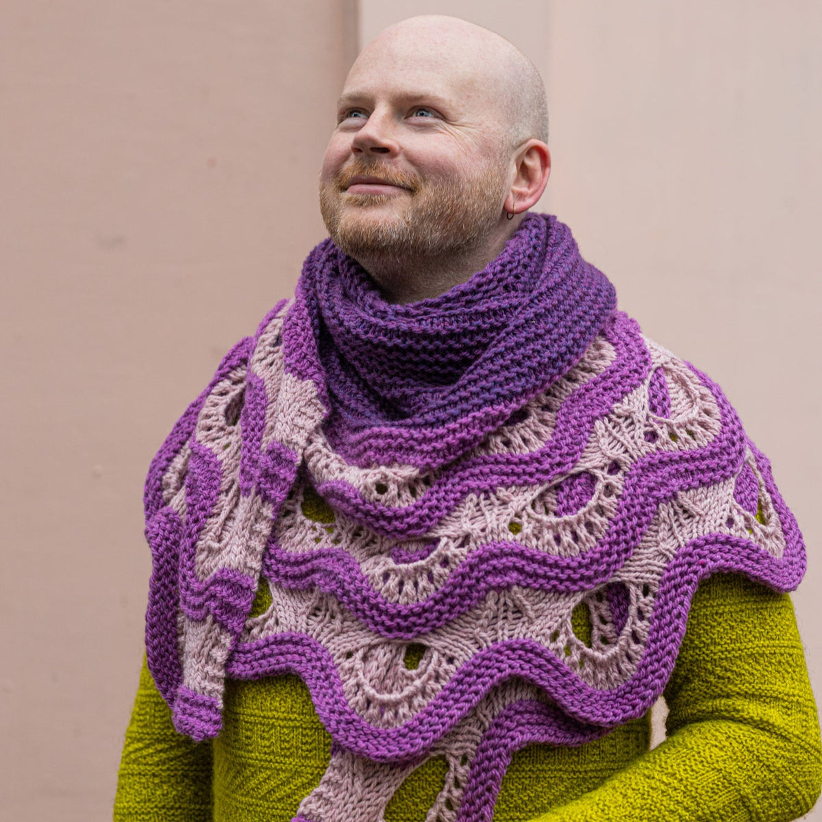 This image shows Stephen West wearing a beautifully knitted sample of the Esjan Shawl in purple and pink tones. He stands in front of a pastel pink background. 