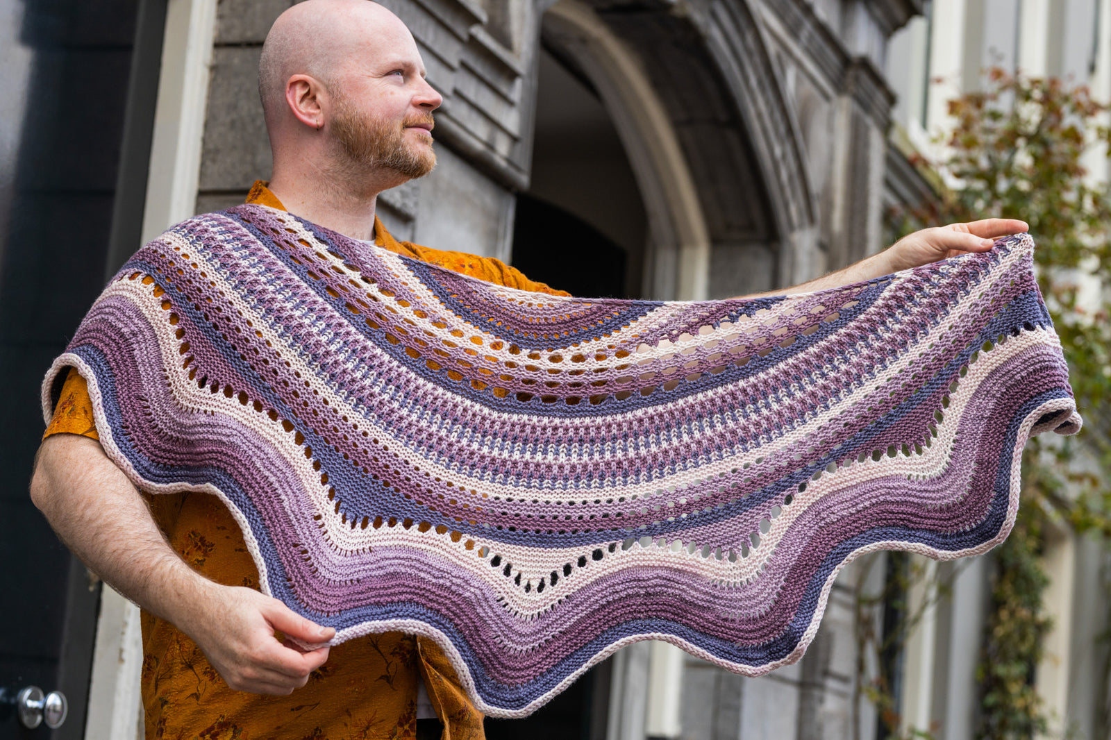 This image shows Stephen West holding a sample of the Eyelet Journey Shawl by Westknits. The sample is knitted in purple and pink gradients. The background shows a building in Amsterdam. 