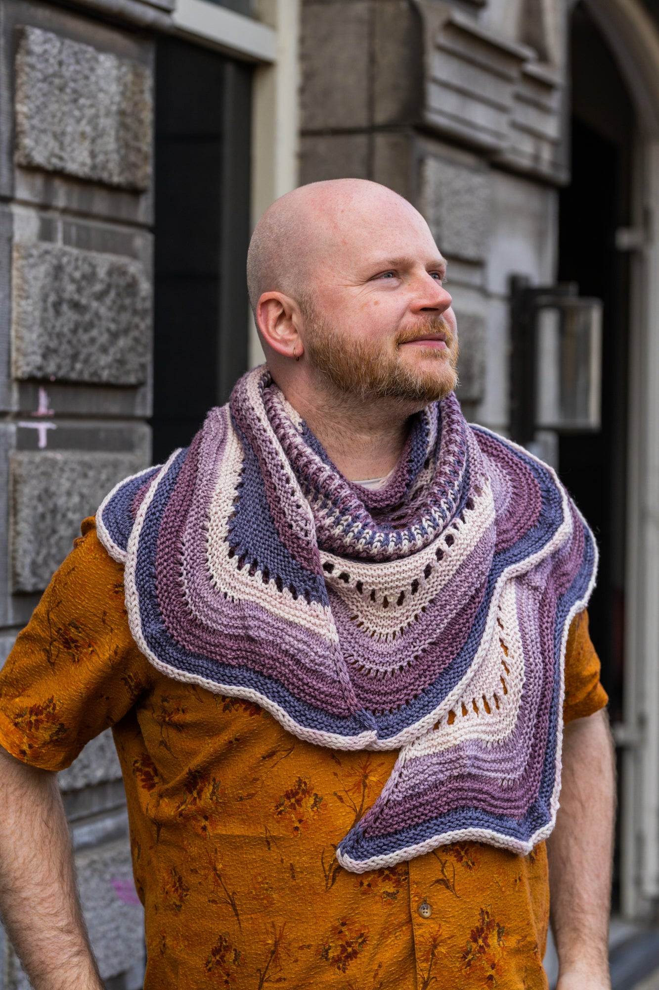 This image shows Stephen West holding a sample of the Eyelet Journey Shawl by Westknits. The sample is knitted in purple and pink gradients. The background shows a building in Amsterdam. 
