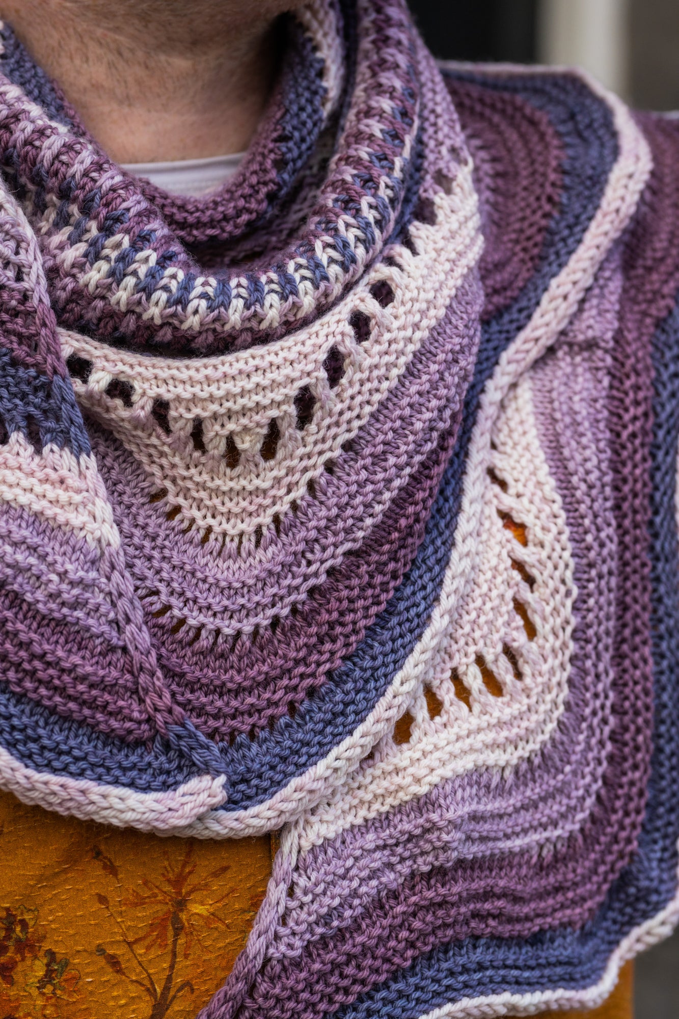 This image shows a close-up of Stephen West holding a sample of the Eyelet Journey Shawl by Westknits. The sample is knitted in purple and pink gradients. The background shows a building in Amsterdam. 