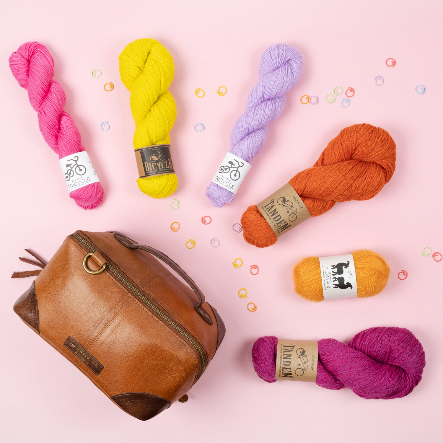 Various skeins of yarn and various notions in bright colours around a brown Re:Designed project bag on a lilac background