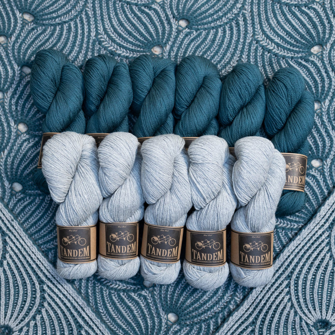 This image shows a detail of a beautifully knitted sample of the Flying Foxtail Blanket by Westknits in grey and blue tones. The yarn in the same colours lays on top. 