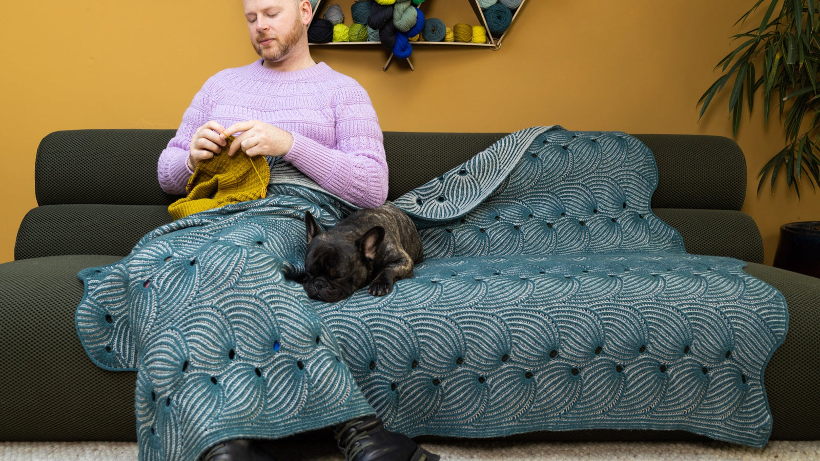 This image shows Stephen West knitting while wearing a beautifully knitted sample of the Flying Foxtail Blanket by Westknits in grey and blue tones. A dog lays on the blanket. 