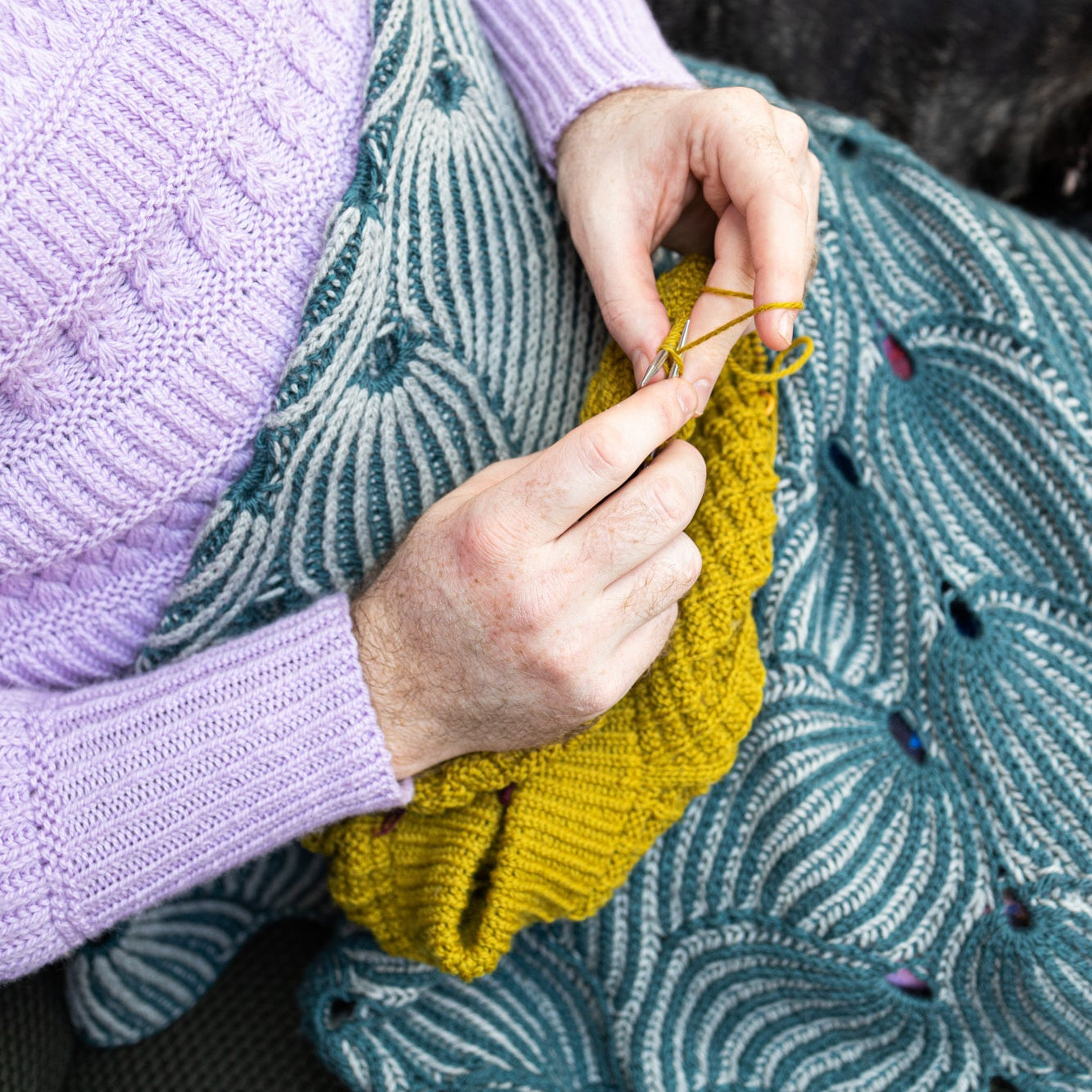 This image shows Stephen West knitting while wearing a beautifully knitted sample of the Flying Foxtail Blanket by Westknits in grey and blue tones. 