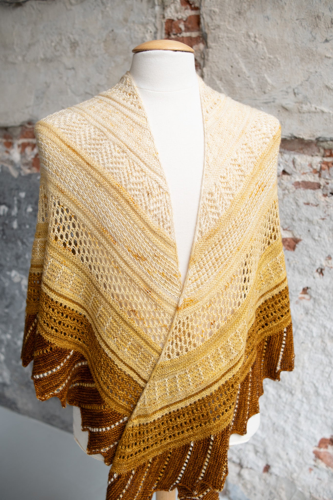 This image shows a sample of the Glittering Snowscape Shawl by Westknits knitted in warm tones. It stands on a brick background. 