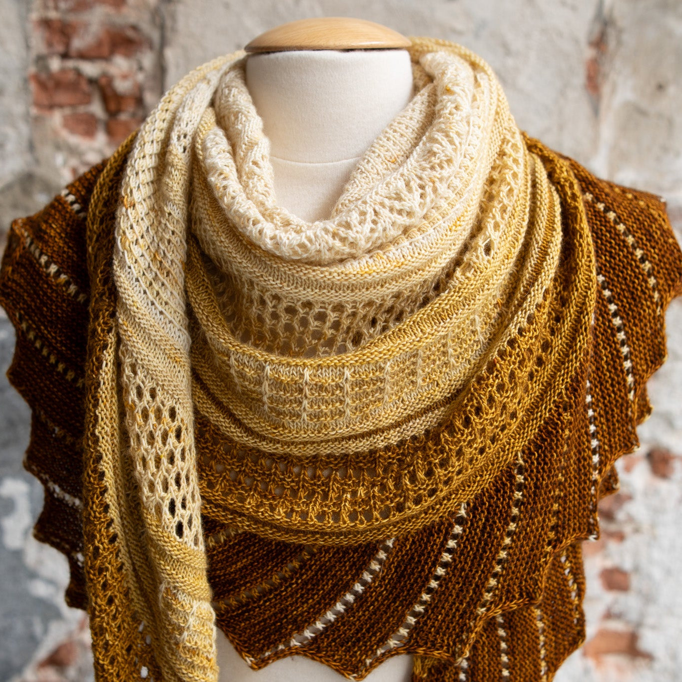 This image shows a sample of the Glittering Snowscape Shawl by Westknits knitted in warm tones. It stands on a brick background. 