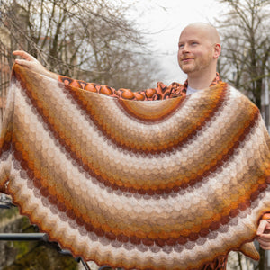 This image shows Stephen West holding a beautifully knitted shawl in warm, earthy tones. The background shows a canal in Amsterdam centre. The shawl is named "The Awesomer Gossamer" and it is launched as part of the Glow Along. 
