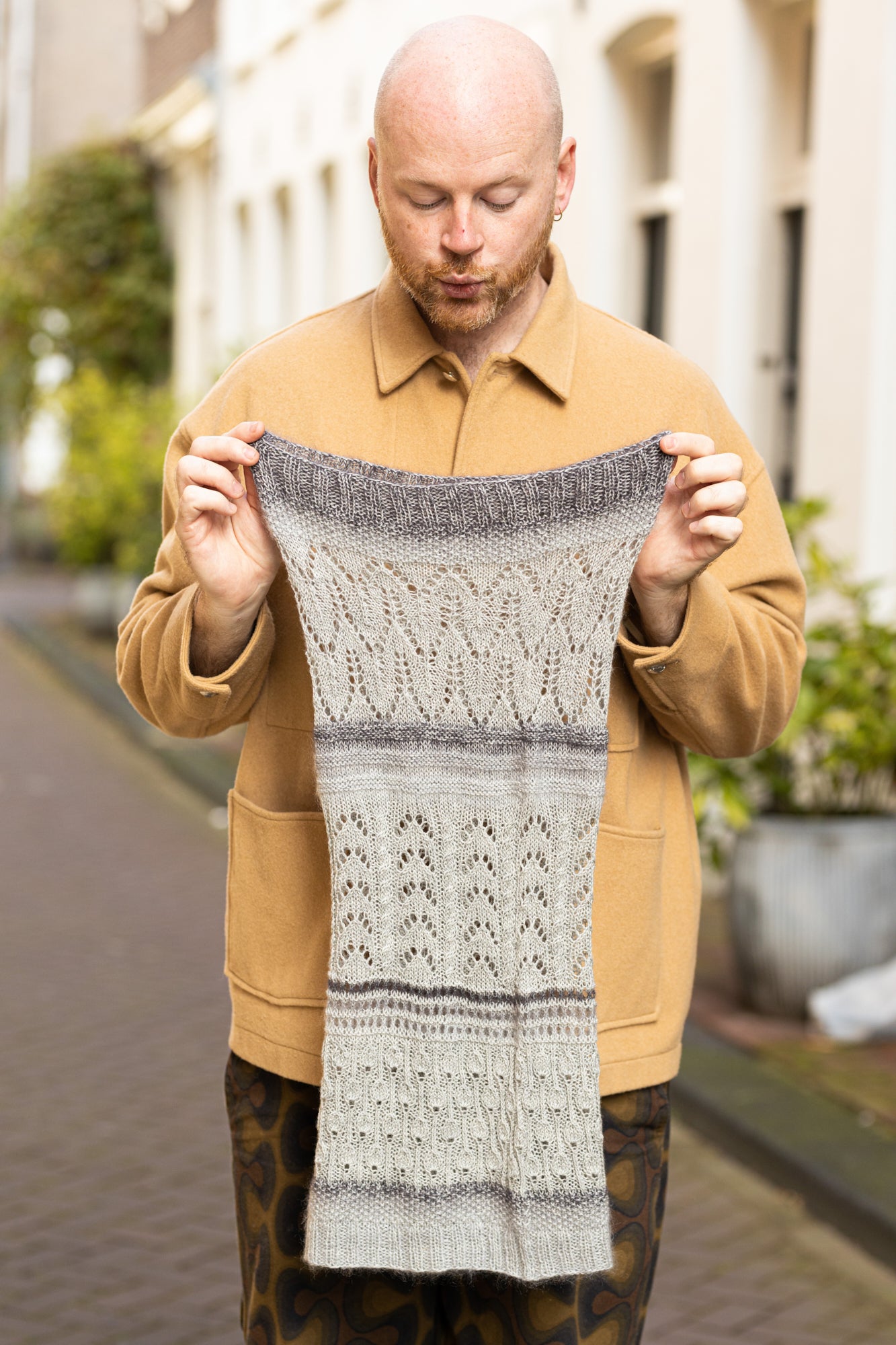Stephen West holding a grey sample of the Glowhair Frost Cowl by Westknits. The background shows a street in Amsterdam. 