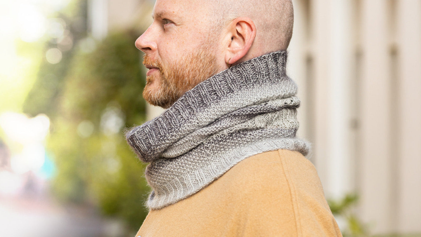 Stephen West Man standing with his profile to camera in a outside blurry setting wearing a handknitted cowl neck scarf in shades of grey
