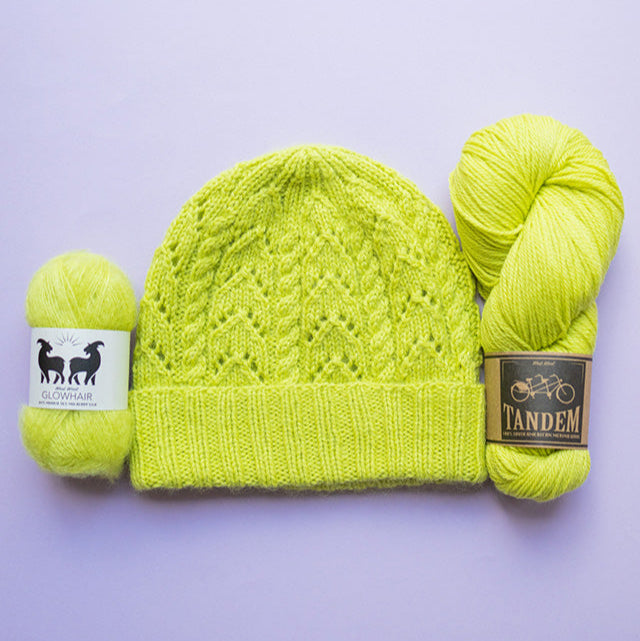 Flat lay image of a knitted beanie hat n green pickle juice colour with a ball kof West Wool mohair and skein of DK merino wool 