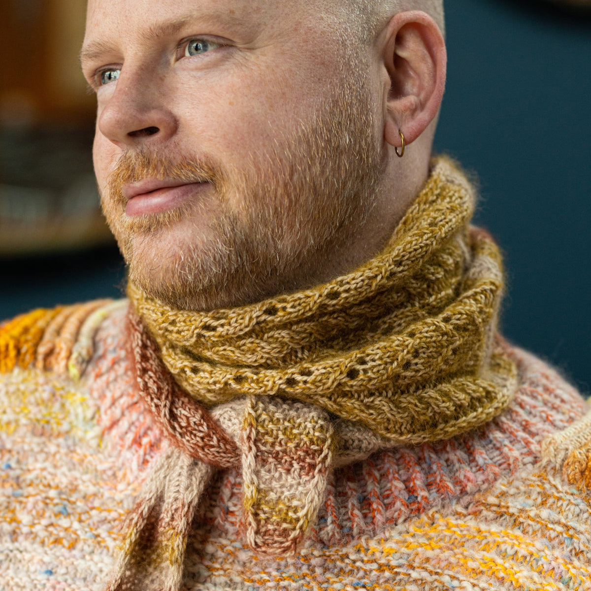 This image shows Stephen West wearing the Glowhair Frost Scarflett. This small shawl is knitted with warm tones, including yellow, cream, and brown. 