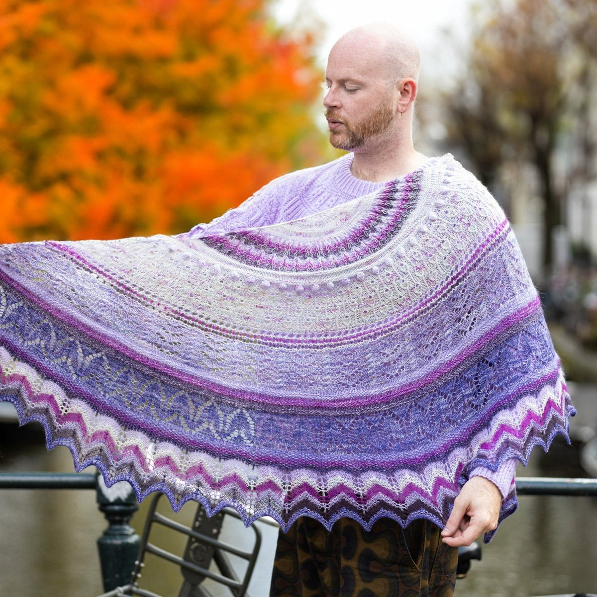 Stephen West holding an intricately knitted shawl in shades of purple, lavender, and frosty grey, featuring lace and textured stitch patterns. The shawl displays a gradient effect with wavy edges, set against a vibrant autumn background with orange foliage and a blurred canal.