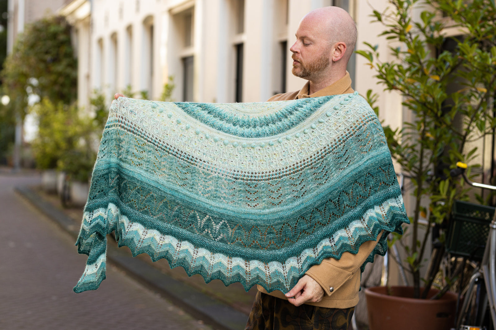 This image shows Stephen West wearing a beautifully knitted sample of the Glowhair Frost Shawl by Westkntis. The sample is knitted in cool shades of white, light blue, and aquamarine. Stephen poses in a street in Amsterdam. 