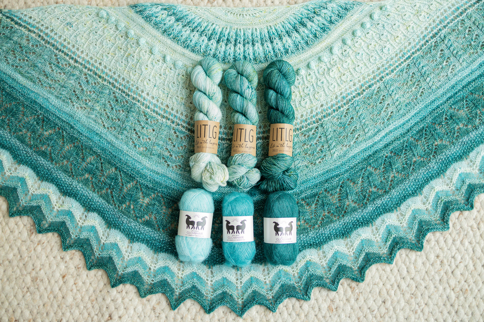 This image shows a beautifully knitted sample of the Glowhair Frost Shawl from Westknits in cool blue and aquamarine tones. The colours of yarn used to knit the sample lay on it. 