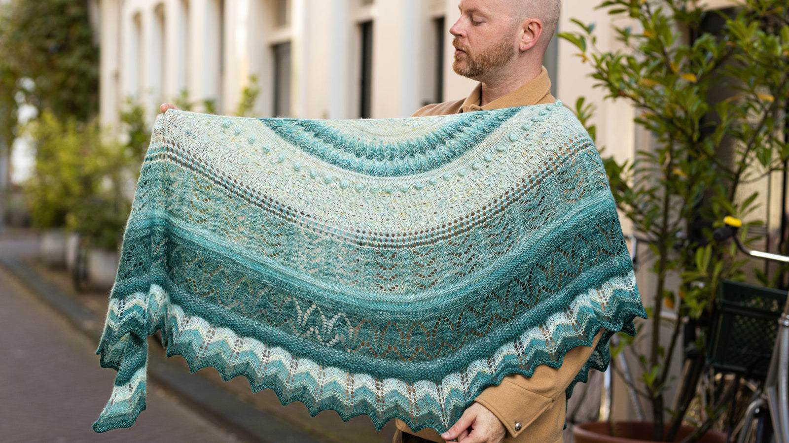 This image shows Stephen West wearing a beautifully knitted sample of the Glowhair Frost Shawl by Westkntis. The sample is knitted in cool shades of white, light blue, and aquamarine. Stephen poses in a street in Amsterdam. 