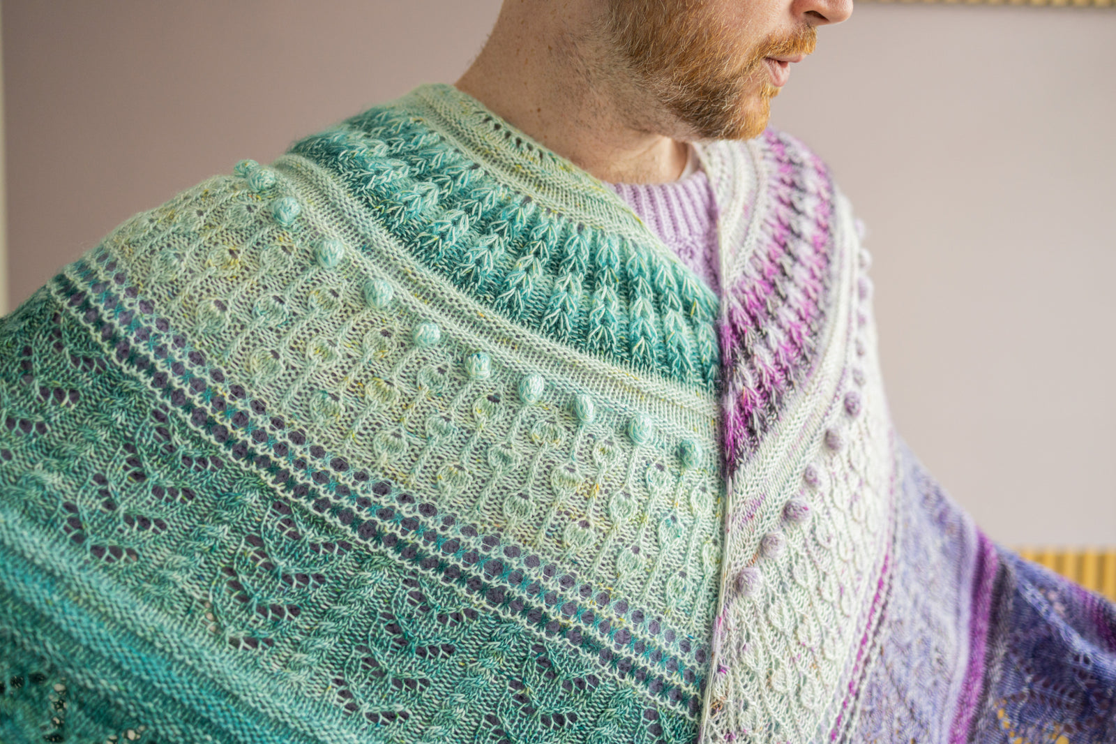 This image shows Stephen West wearing two beautifully knitted sample of the Glowhair Frost Shawl by Westkntis. The sample are knitted in cool green shades and purple tones. 