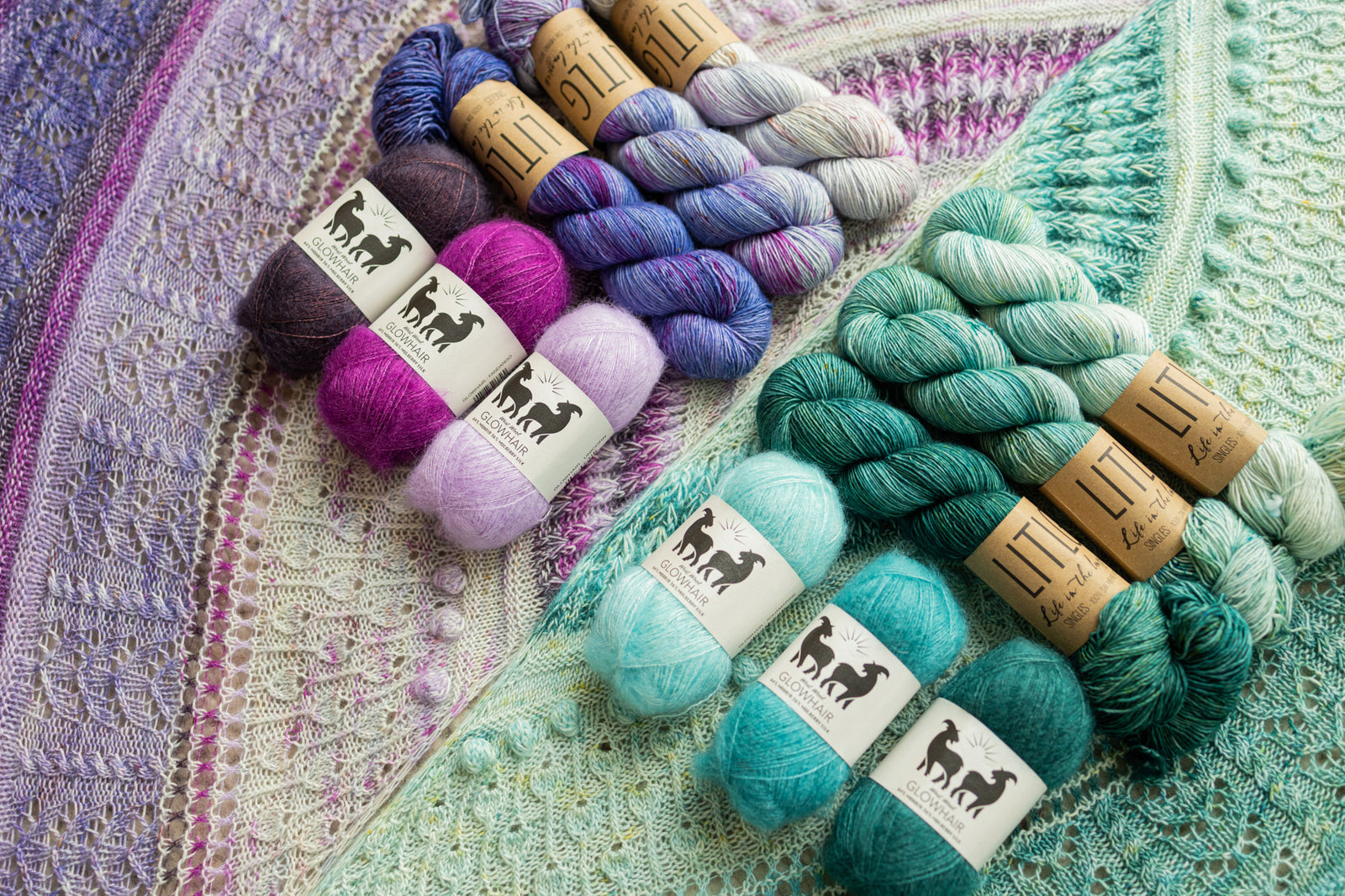 This image shows 2 beautifully knitted sample of the Glowhair Frost Shawl from Westknits in cool blue and purple tones. The colours of yarn used to knit the sample lay on it. 