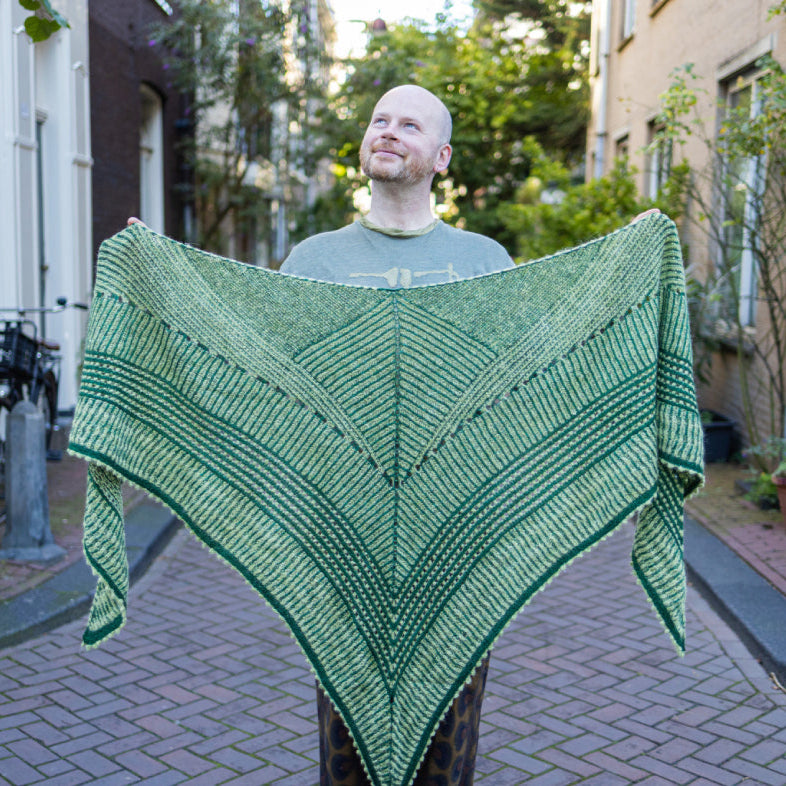 This image shows Stephen West holding the Húsafell Shawl. The shawl is knitted with green tones. The background shows a street in Amsterdam centre. 