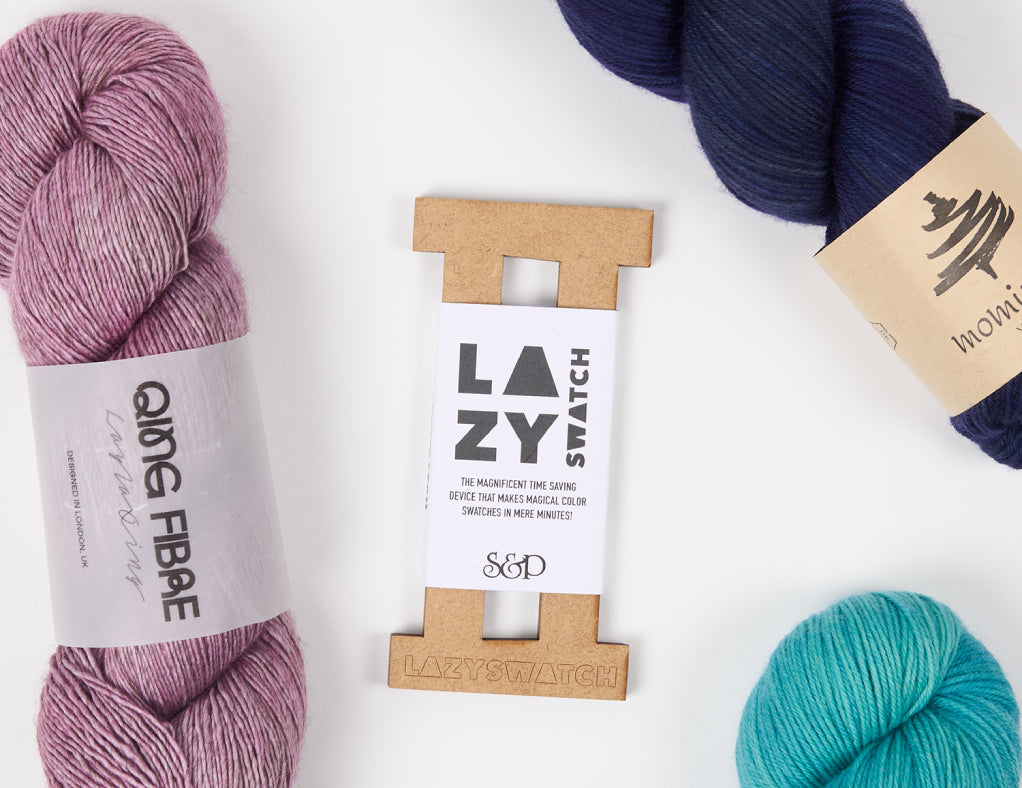 This image shows a Lazy Swatch from Stephen & Penelope surrounded by three skeins of yarn in blue and pink tones. They stand on a white background. 