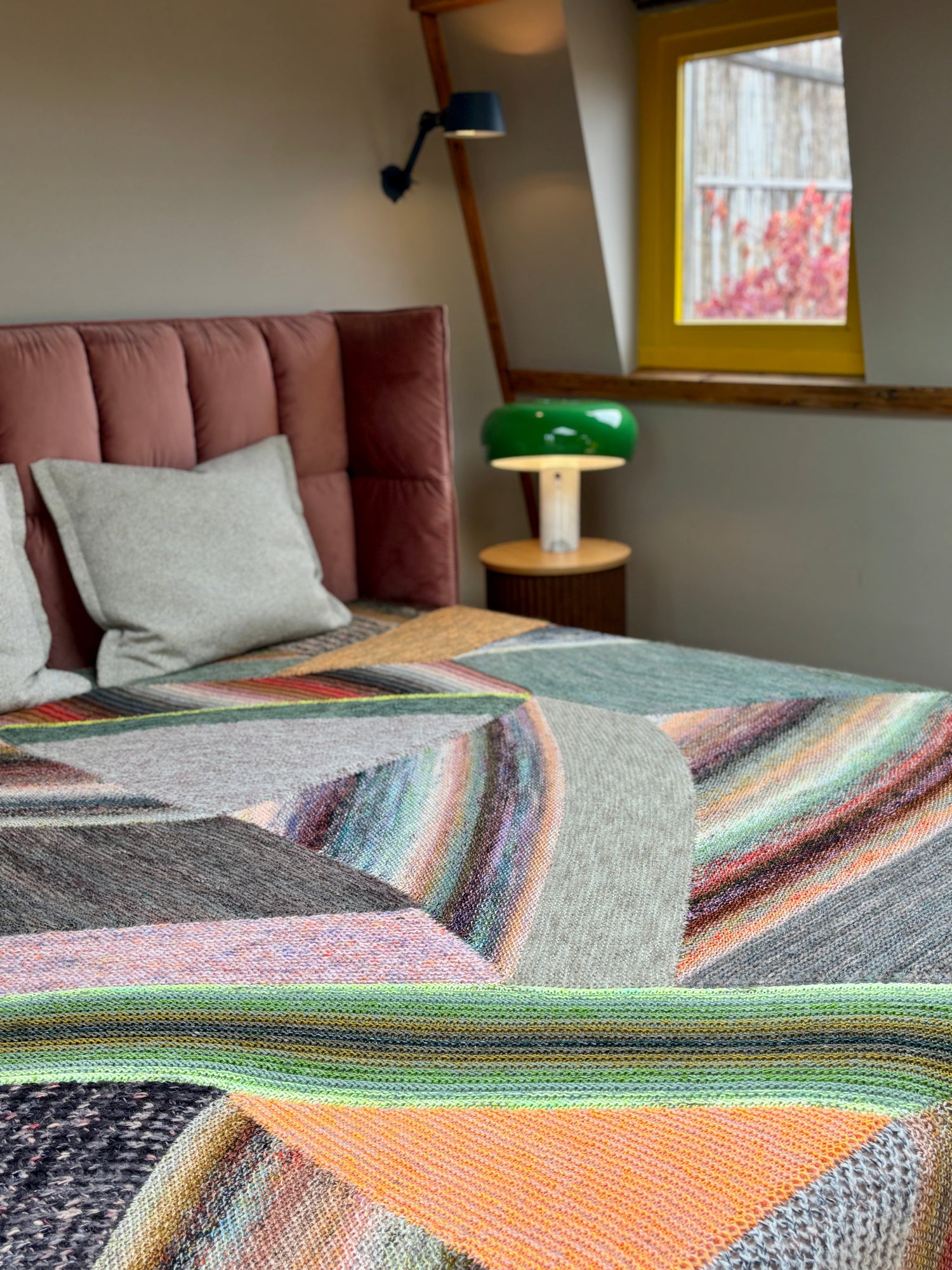 This image shows a beautifully knitted sample of the Marled Magic Blanket by Westknits. The blanket is laid on a bed and it is knitted with a patchwork layout in various colours. 