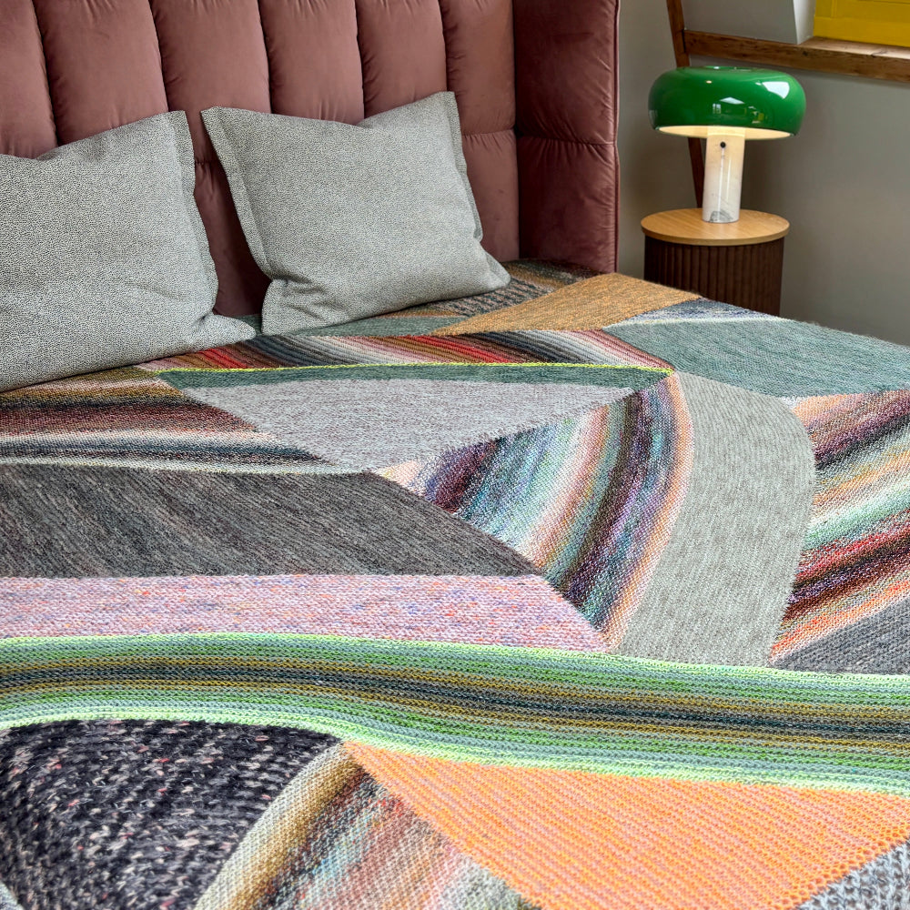 This image shows a beautifully knitted sample of the Marled Magic Blanket by Westknits. The blanket is laid on a bed and it is knitted with a patchwork layout in various colours. 